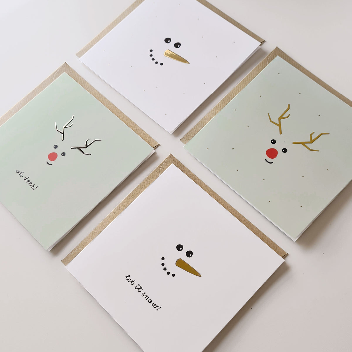 Afbeelding van 4 verschillende kerstkaarten met goudfolie, liggend op kraft enveloppen.