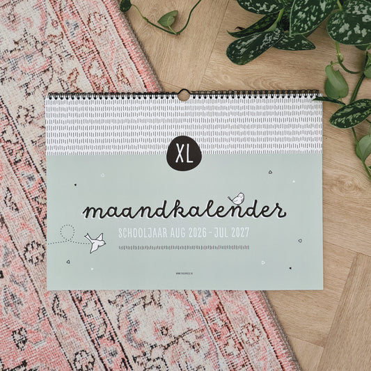 Sfeerbeeld XL maandkalender schooljaar 2026 2027 op roze Marrokaans vloerkleed met plant ernaast.