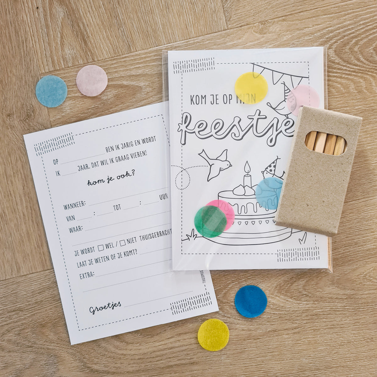 Sfeerbeeld van de uitnodigingskaarten ingepakt zoals je ze ook kunt bestellen met transparant zakje en een doosje kleurpotloodjes. op houten tafel met confetti.