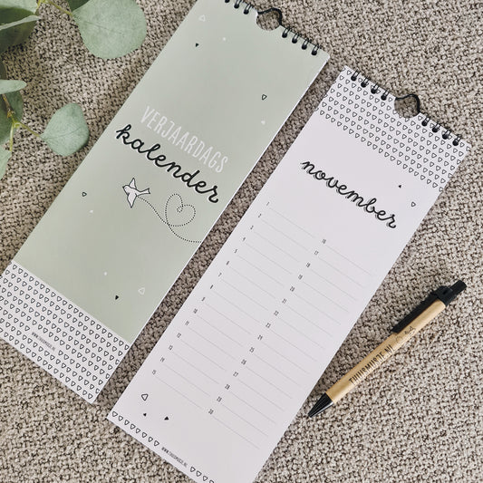 Sfeerfoto van voorkant kalender en pagina november. Ernaast een balpen welke de grootte van de kalender laat zien, welke 10,5x30cm is.