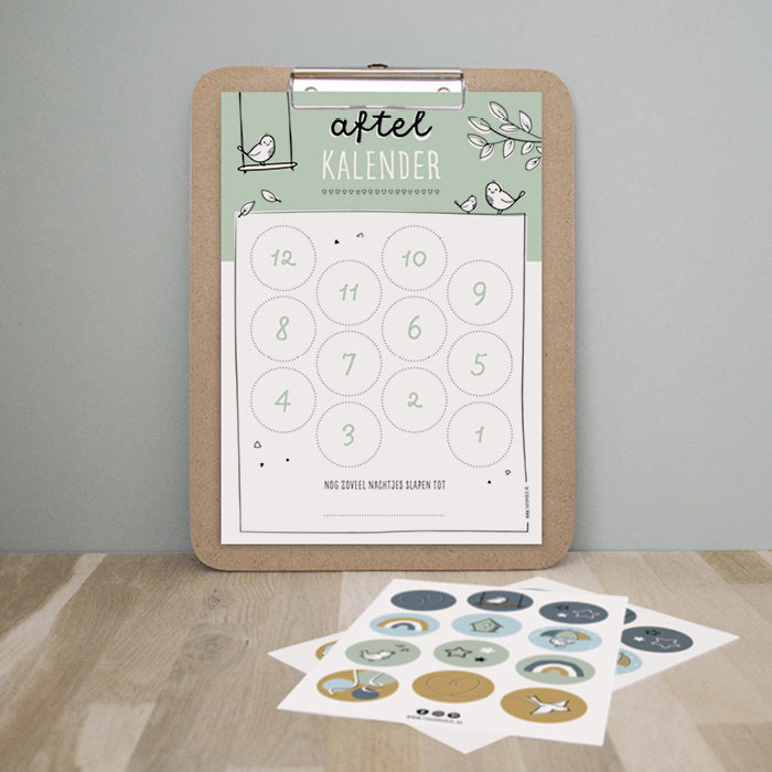 Aftelkalender groen hangend op klembord met ervoor op tafel het kleurrijke stickervel. 