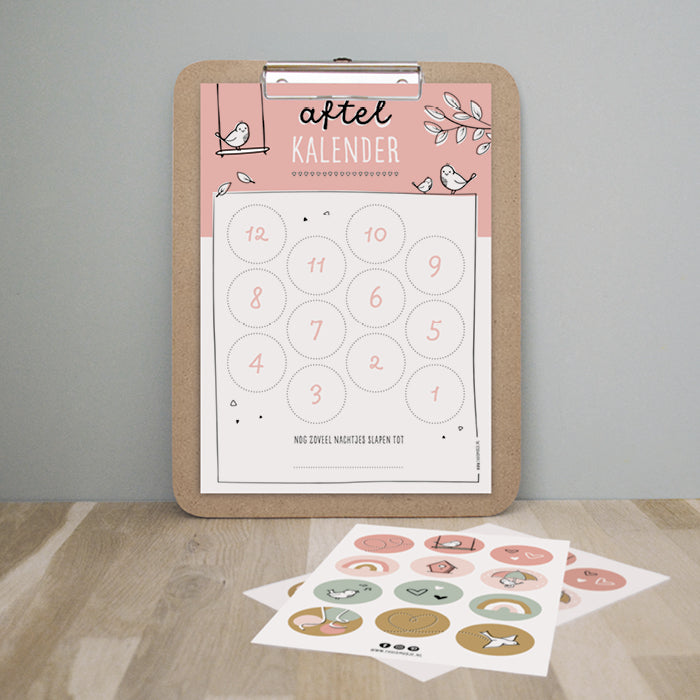 Aftelkalender roze hangend op klembord met ervoor op tafel het kleurrijke stickervel. 