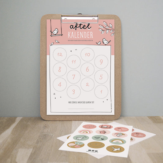 Aftelkalender roze hangend op klembord met ervoor op tafel het kleurrijke stickervel. 