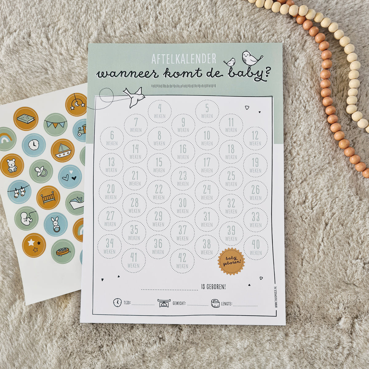 Sfeerbeeld van aftelposter zwangerschap op zacht kleed met kralenketting en stickervel. 