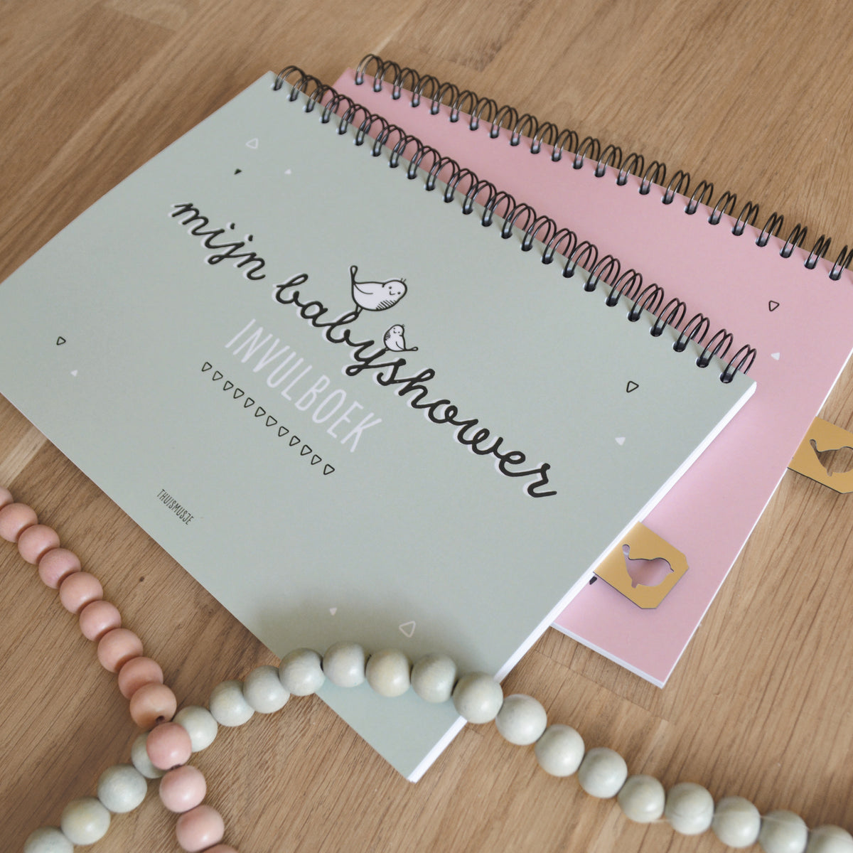 Sfeerfoto van roze en groene babyshower invulboeken op een houten tafel met roze en groene kralenkettingen erbij. 