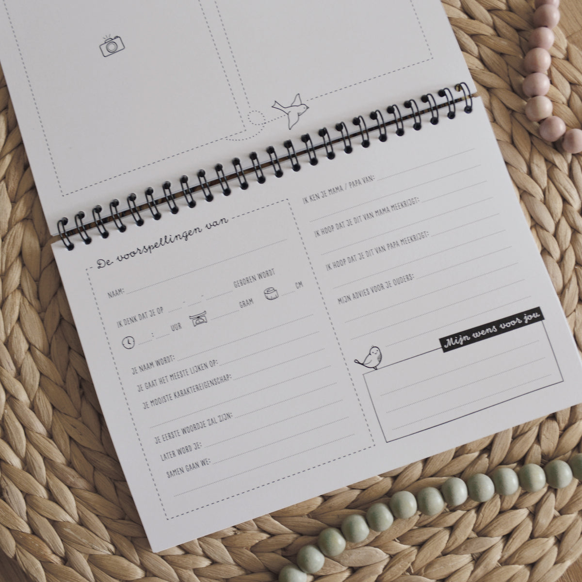Sfeerbeeld met inkijkje in babyshower invulboek. De titel is de voorspellingen van± waarbij iedere gast voorspellingen kan doen en vragen kan beantwoorden. Sfeervol beeld op geweven ondergrond met roze en groene kralenketting.