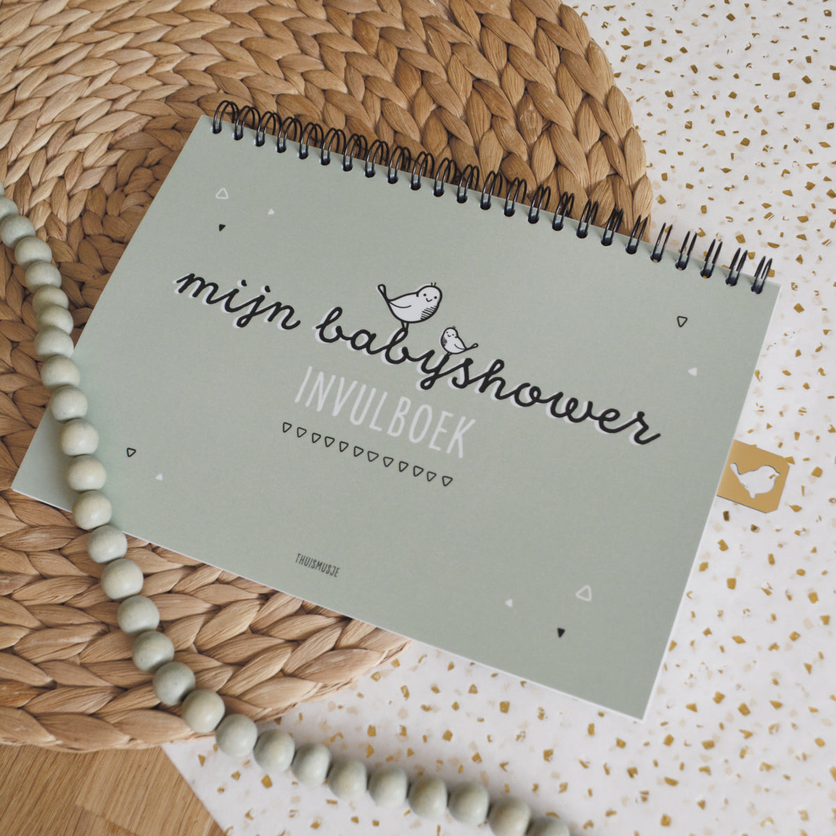 Sfeerbeeld van mijn babyshower invulboek op een met riet geweven ondergrond, vrolijk inpakpapier en een groene kralenketting. 