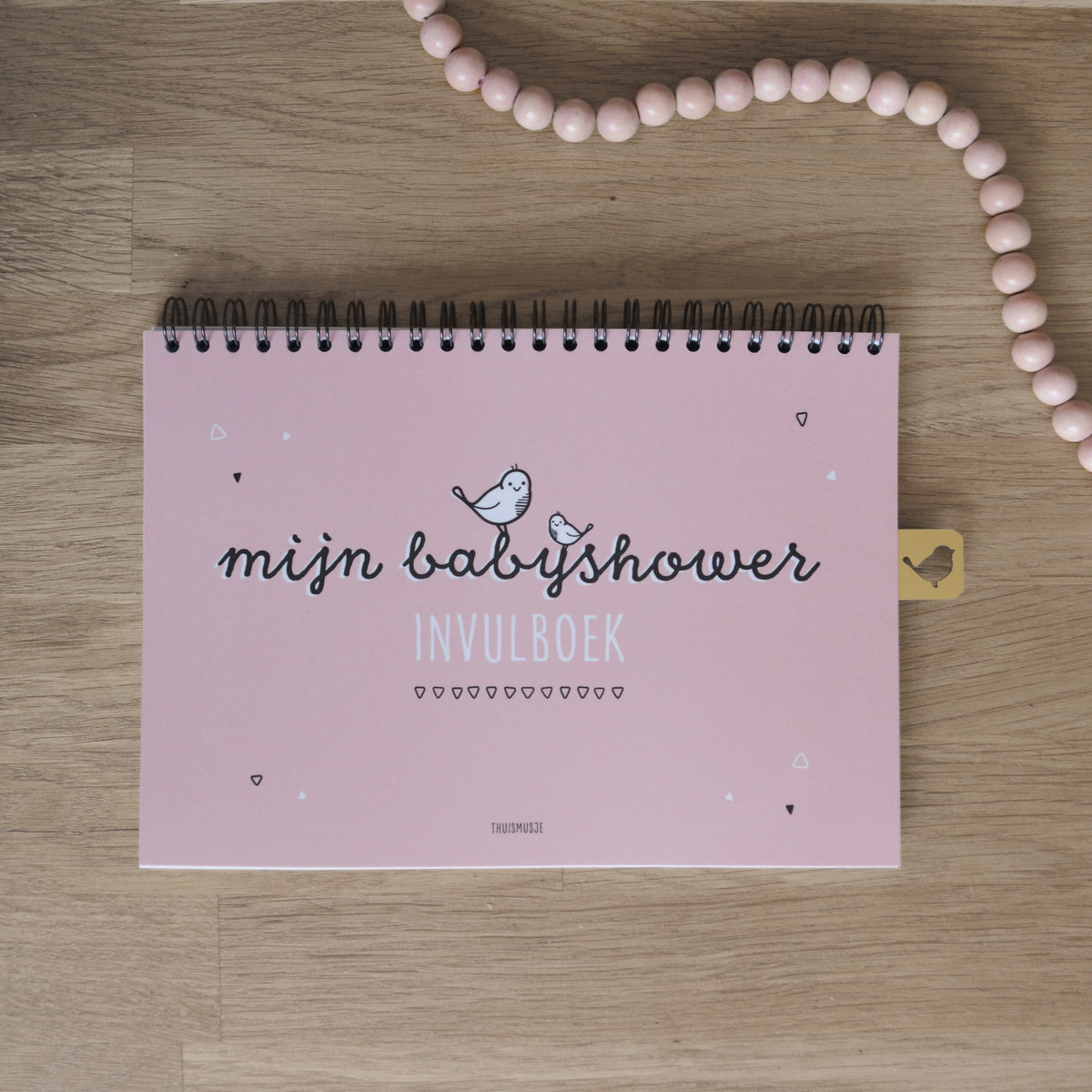 Sfeerbeeld van mijn babyshower invulboek op houten tafel met een roze kralenketting. 