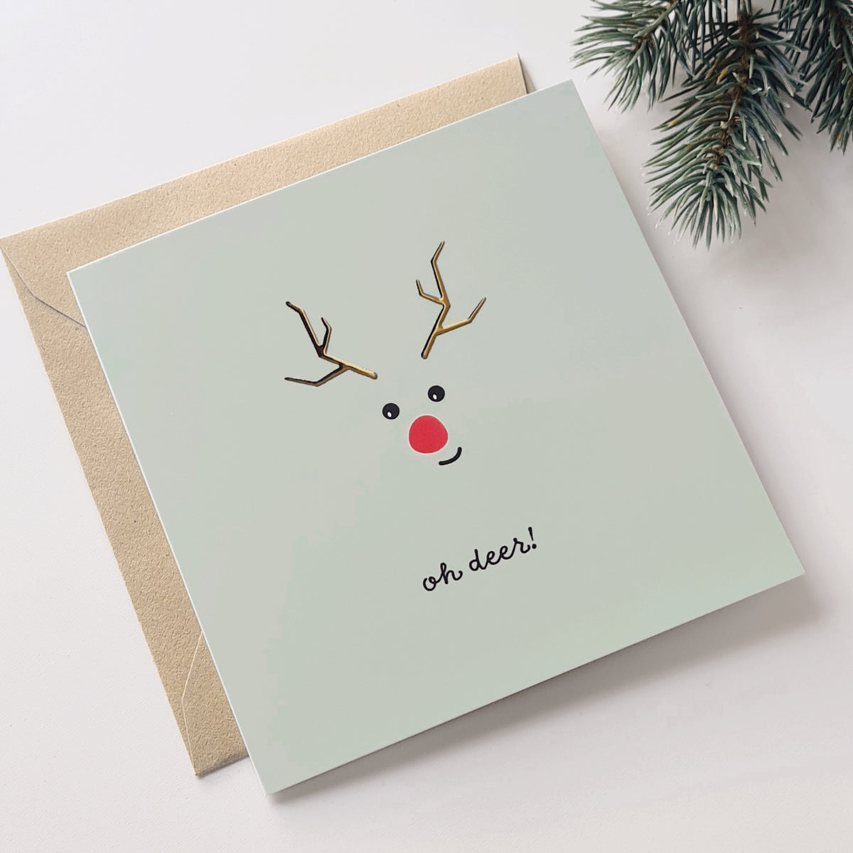 kerstkaart met rendiertje met goudfolie erop en de tekst: oh deer! weergegeven met kraft envelop en een dennentak