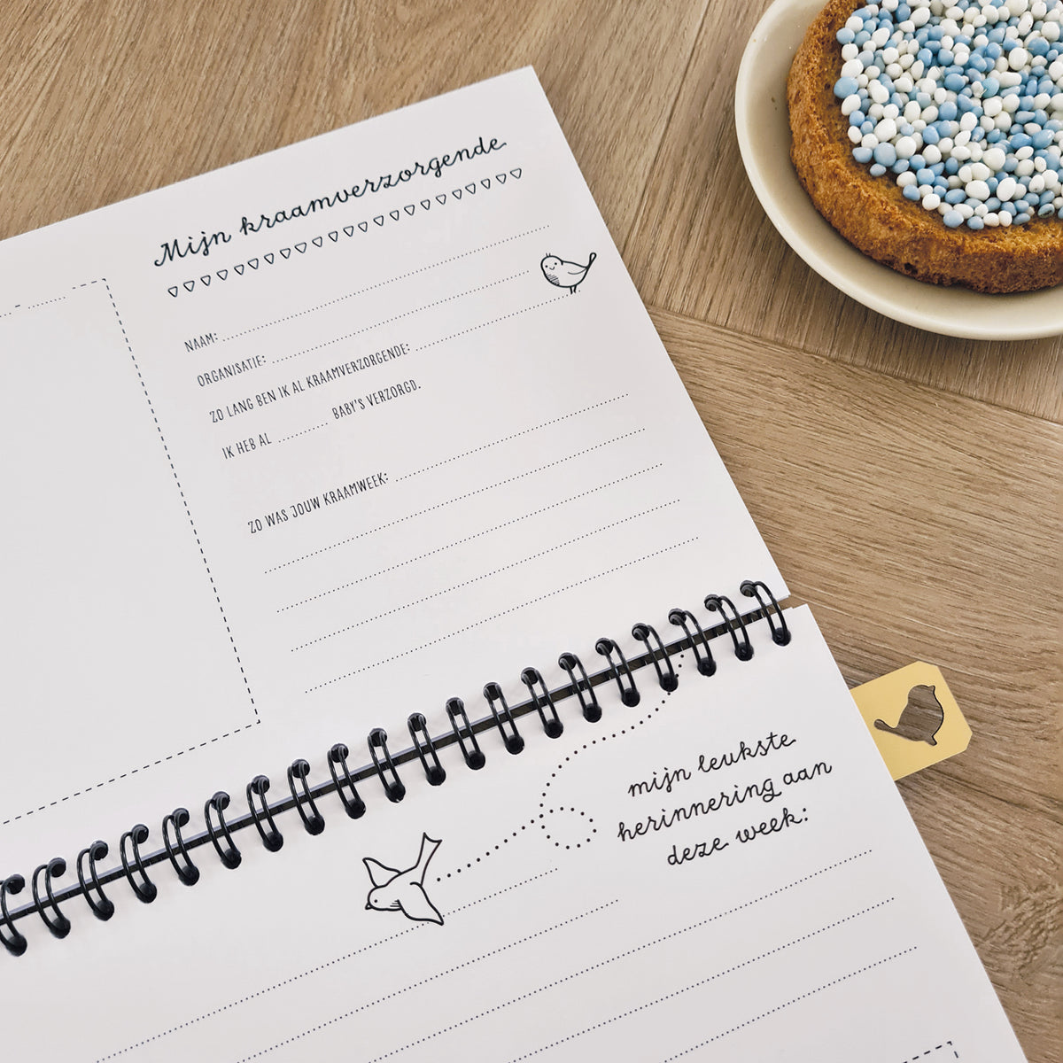 Sfeerbeeld van kraambezoek invulboek op houten ondergrond met beschuit met blauwe muisjes. Inkijkje in boek op pagina voor de kraamverzorgende. 