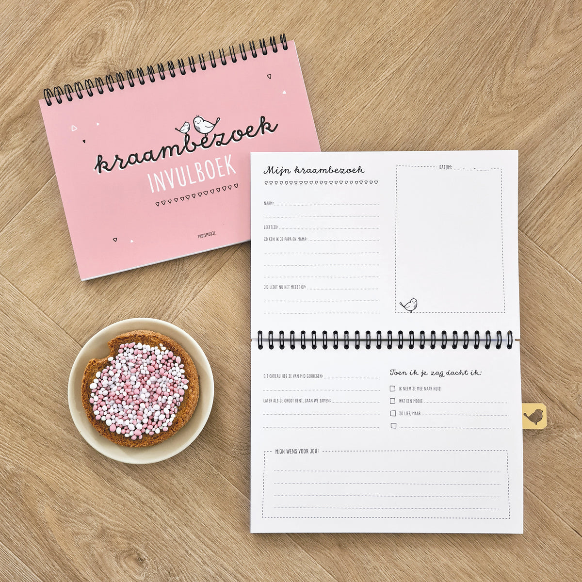 Sfeerbeeld roze kraambezoekboek op houten achtergrond. Opengeslagen boek geeft inkijkje in binnenzijde met invulbare vragen voor het kraambezoek en ruimte voor een foto. Op tafel staat een beschuit met muisjes.