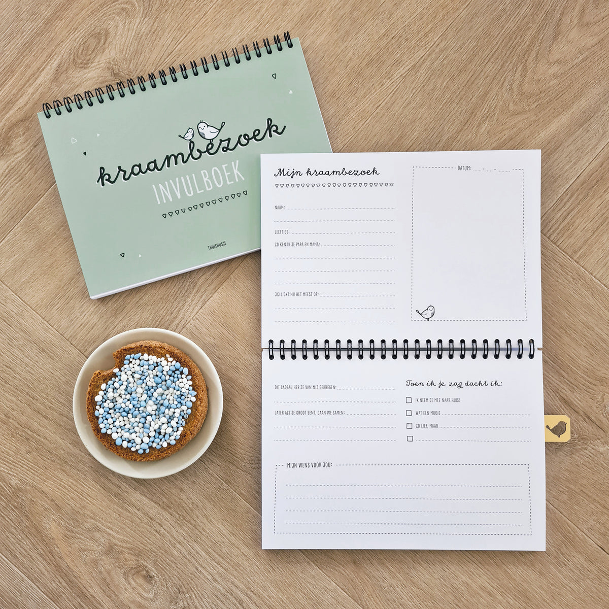 Sfeerbeeld groene kraambezoekboek op houten achtergrond. Opengeslagen boek geeft inkijkje in binnenzijde met invulbare vragen voor het kraambezoek en ruimte voor een foto. Op tafel staat een beschuit met muisjes.