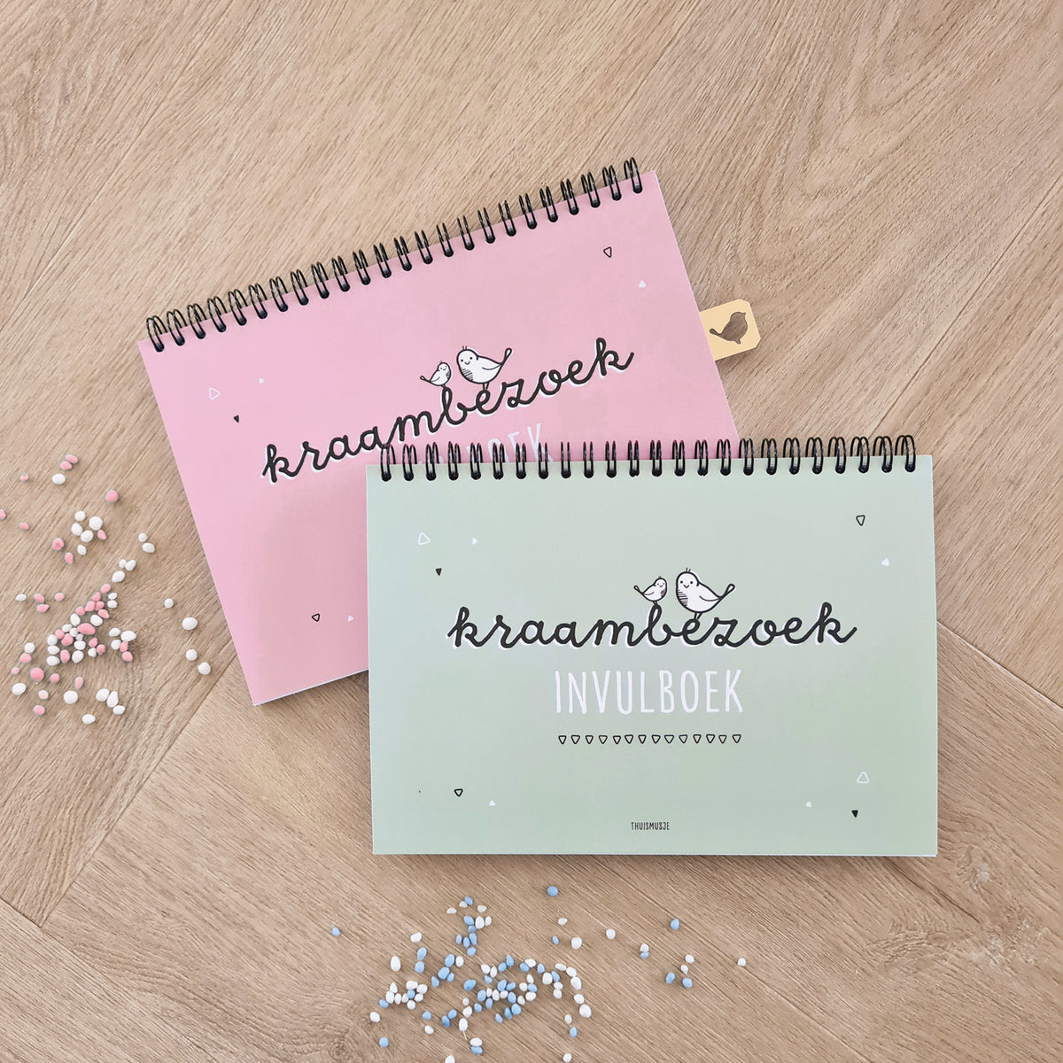 Sfeerbeeld van roze en groen kraambezoek invulboek op houten ondergrond met rondom gestrooide roze en blauwe muisjes. 