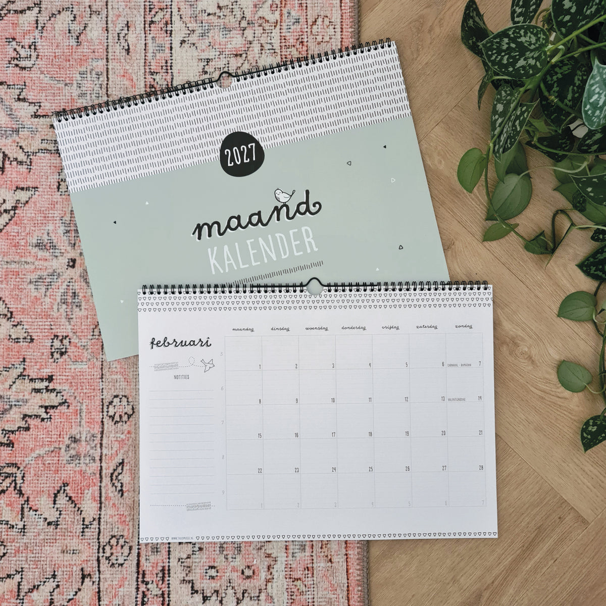 Sfeerbeeld van twee maandkalenders 2027 op vloerkleed met plant ernaast. Gesloten kalender met daarboven een omgeslagen kalender op februari. 
