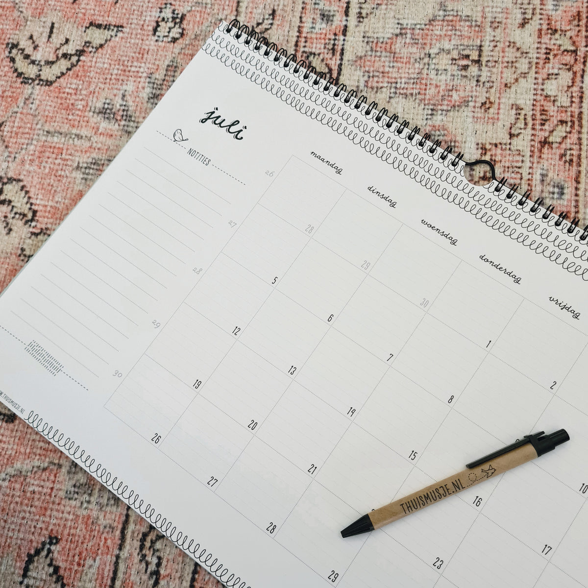Detailfoto van de A3 maandkalender op de maand juli. Liggend op roze vloerkleed met balpen ter indicatie van grootte. 