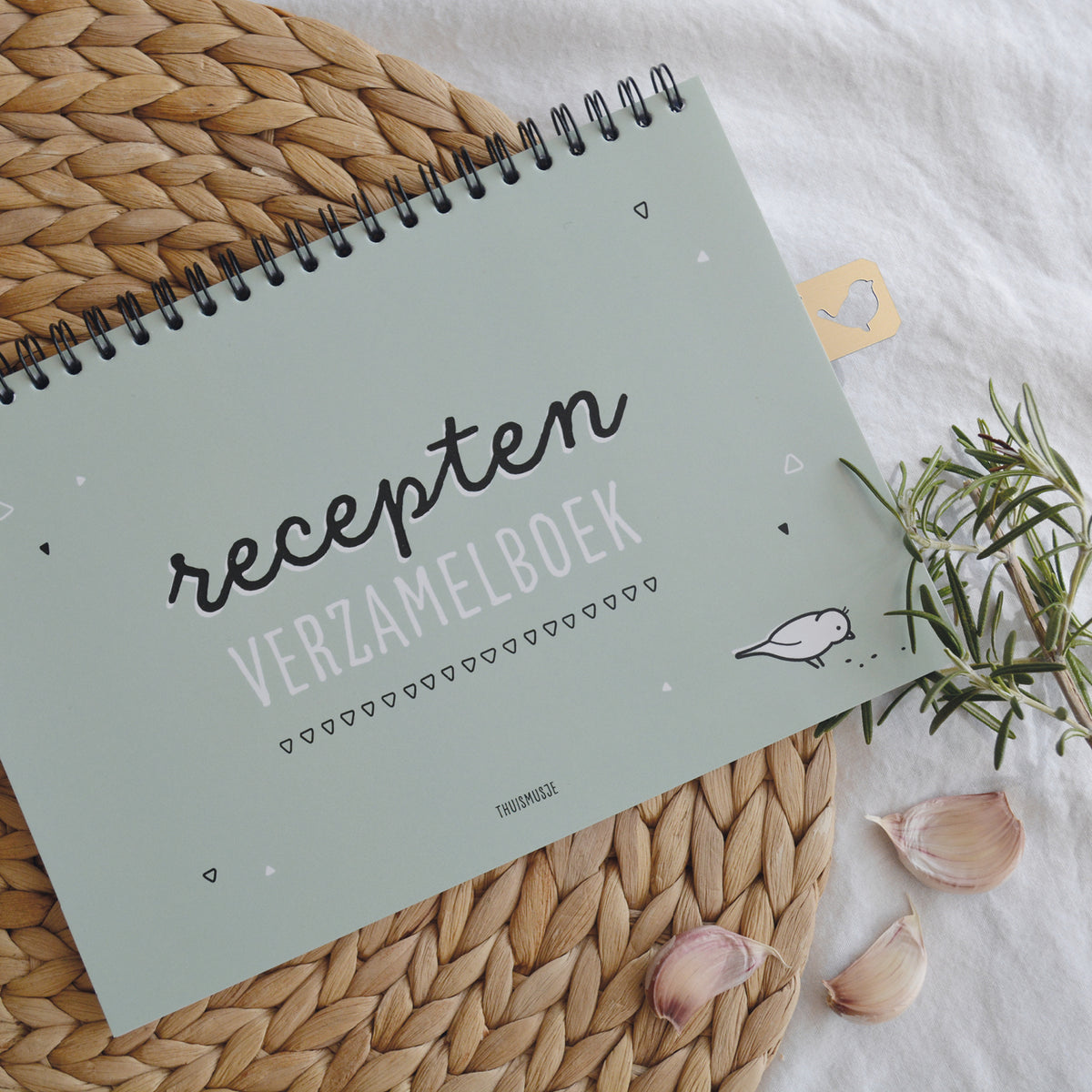 Sfeerfoto van dicht receptenboek op tafelkleed met gouden bladwijzer, takje rozemarijn en knoflooktenen.
