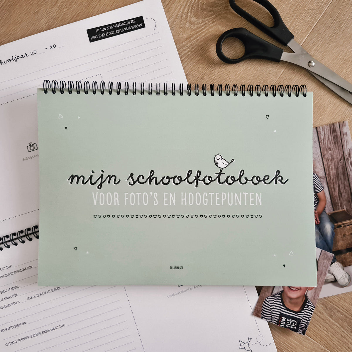 Sfeerbeeld vooraanzicht Mijn schoolfotoboek voor foto's en hoogtepunten. Liggend op houten tafel met schaar, schoolfoto's en opengeslagen schoolfotoboek. 