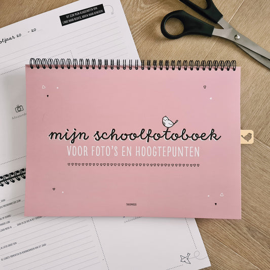 Sfeerbeeld van roze schoolfotoboek op houten tafel met schaar en opengeslagen schoolfotoboek. 
