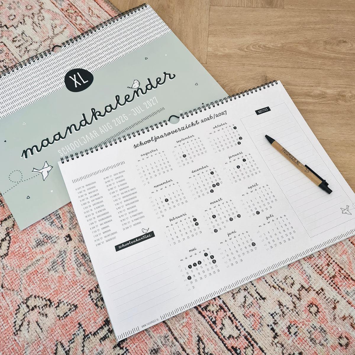Sfeerfoto van 2 A3 kalenders op roze vloerkleed met pen ter indicatie van grootte. Kalender ligt opengevouwen op het schooljaaroverzicht met alle maanden en feestdagen in een duidelijk overzicht.  