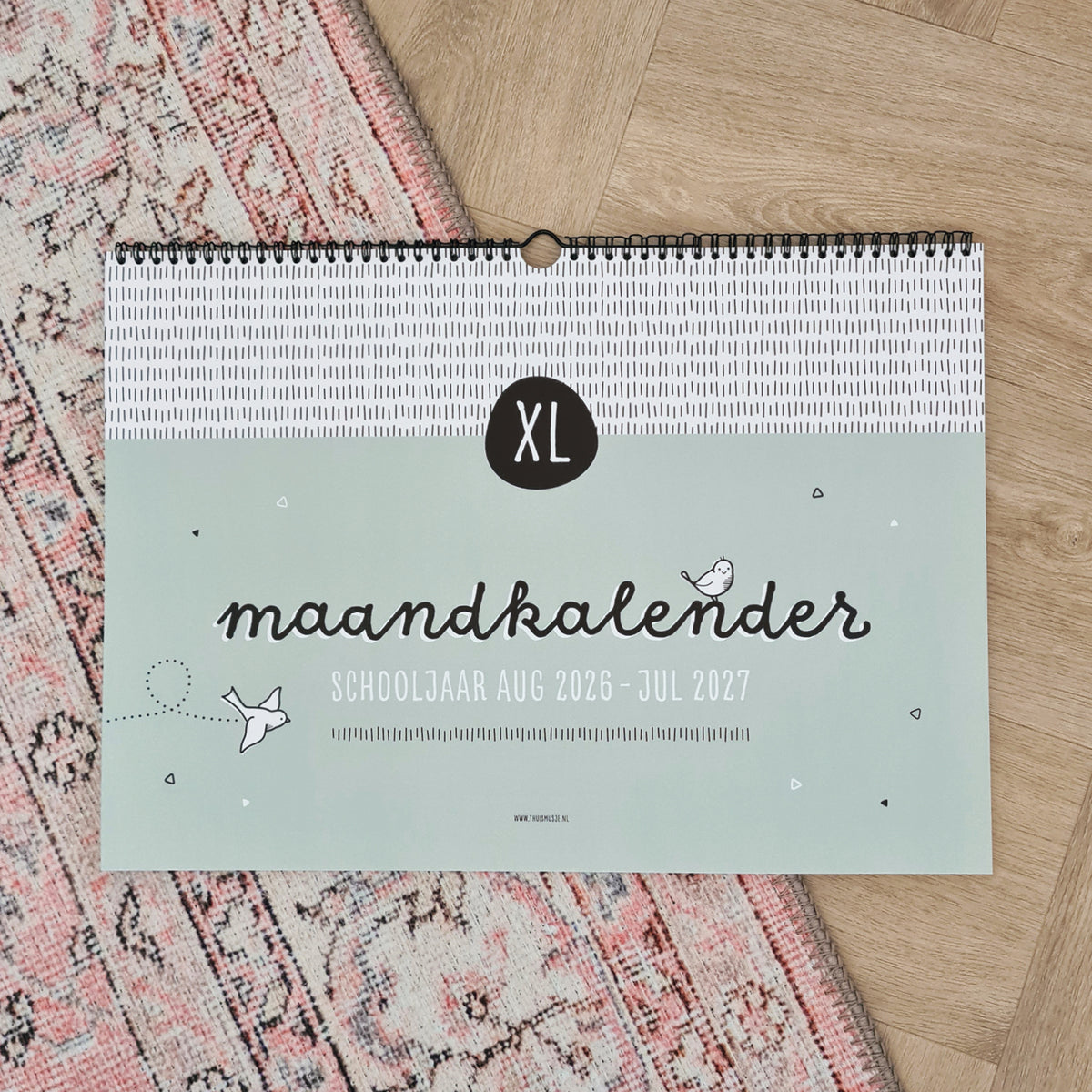 Sfeerfoto vooraanzicht XL maandkalender schooljaar augustus 2026 tot en met juli 2027. Liggend op roze kleed en houten visgraat vloer. 