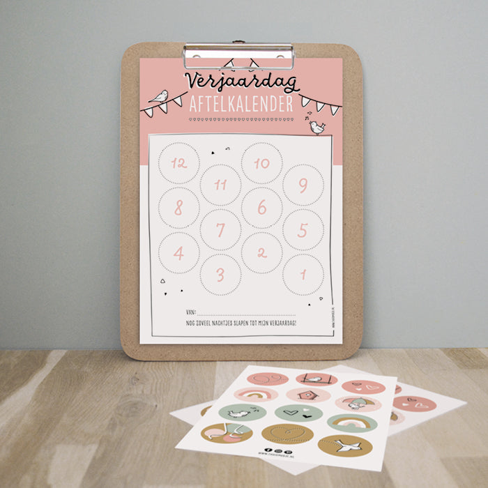 Roze A4 verjaardagsaftelkalender op een klembord met voor op tafel het kleurrijke stickervel met 12 stickers.