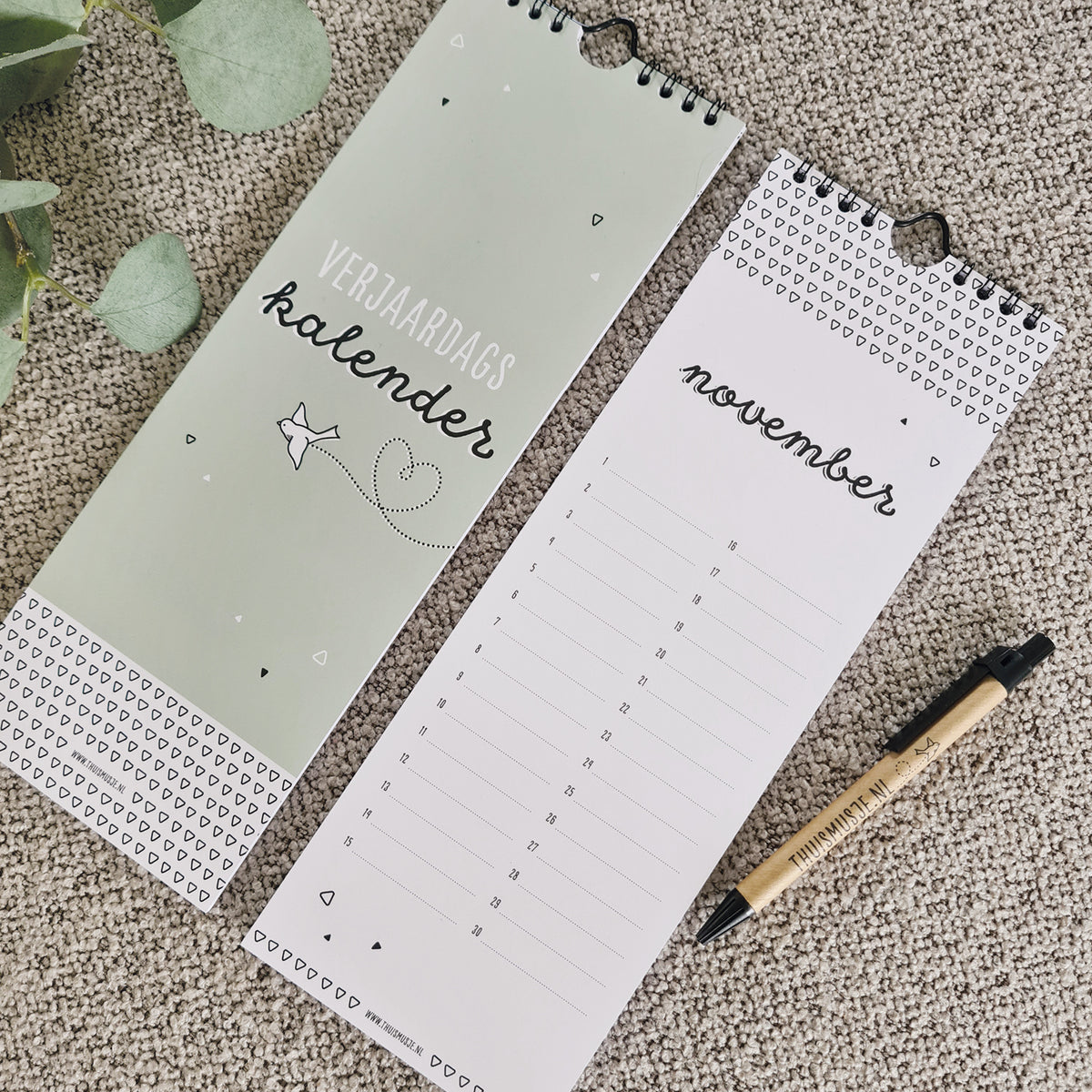 Sfeerfoto van voorkant kalender en pagina november. Ernaast een balpen welke de grootte van de kalender laat zien, welke 10,5x30cm is. 