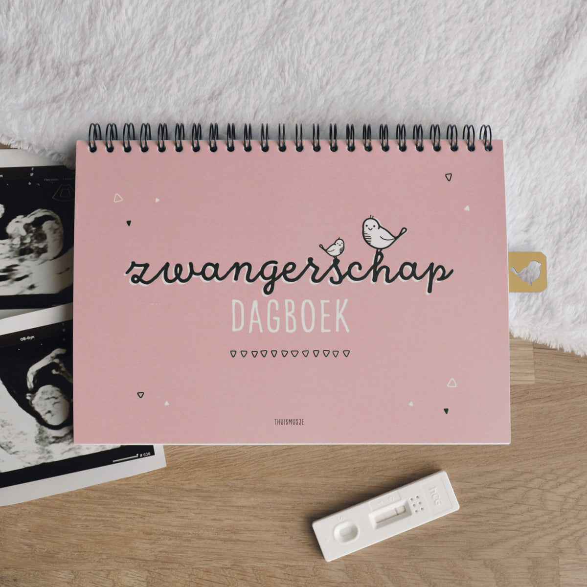 Sfeerbeeld van roze zwangerschapsdagboek. Gesloten boek op houten tafel met zacht kleedje, zwangerschapstest en echofoto´s. 
